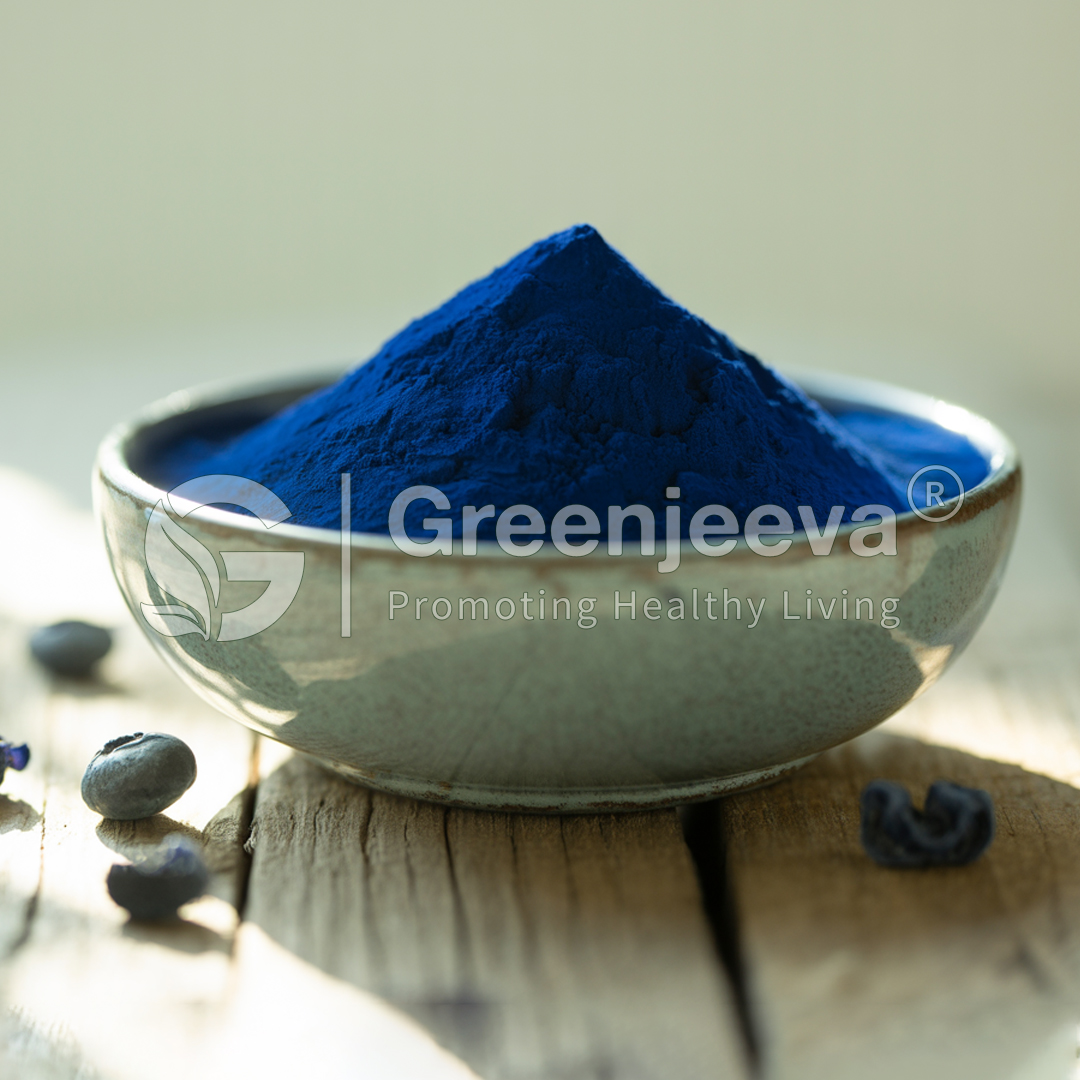 A bowl of vibrant blue powder, surrounded by dark blue berries, placed on a rustic wooden surface.